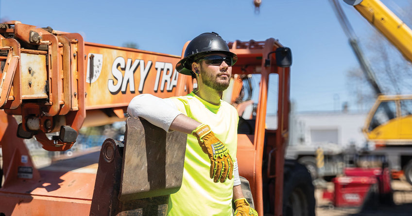 Construction worker standing in front of heavy machinery wearing HexArmor's full brim safety helmet and work gloves.